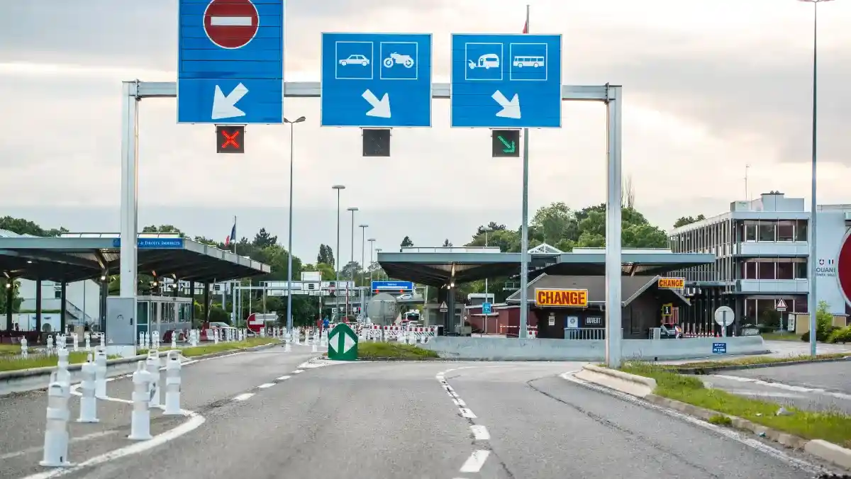 Border checkpoint with lanes for different vehicle types, marked by signs above; a closed lane and open lane indicated. Booths and greenery in the background.