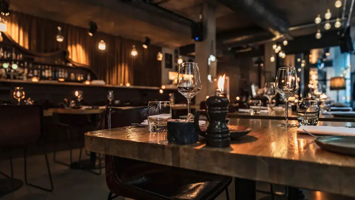 Cozy restaurant interior with dim lighting and elegant decor. Wooden tables are set with wine glasses, candles, and a pepper grinder, exuding a warm, inviting ambiance.