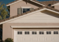 A suburban house with beige walls, a white garage door, and a triangular roof. A tree is on the left, and the sky is clear blue, creating a calm atmosphere.