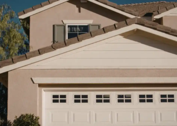 A suburban house with beige walls, a white garage door, and a triangular roof. A tree is on the left, and the sky is clear blue, creating a calm atmosphere.