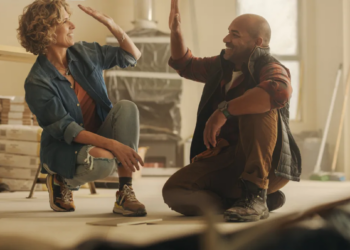 A woman and a man high-five in a warmly lit room under renovation. They smile, conveying teamwork and accomplishment. Stacks of wood are visible in the background.