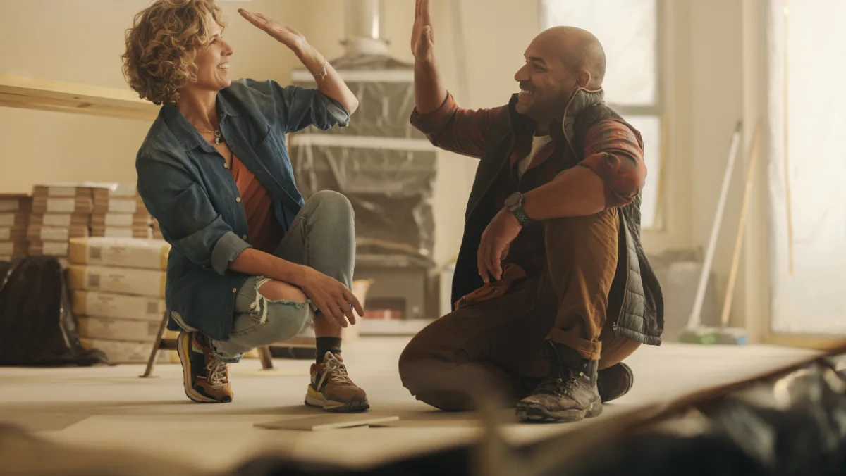 A woman and a man high-five in a warmly lit room under renovation. They smile, conveying teamwork and accomplishment. Stacks of wood are visible in the background.
