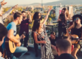Group of friends enjoying a rooftop party, with people singing and playing guitars. Energetic atmosphere with a cityscape backdrop at sunset.