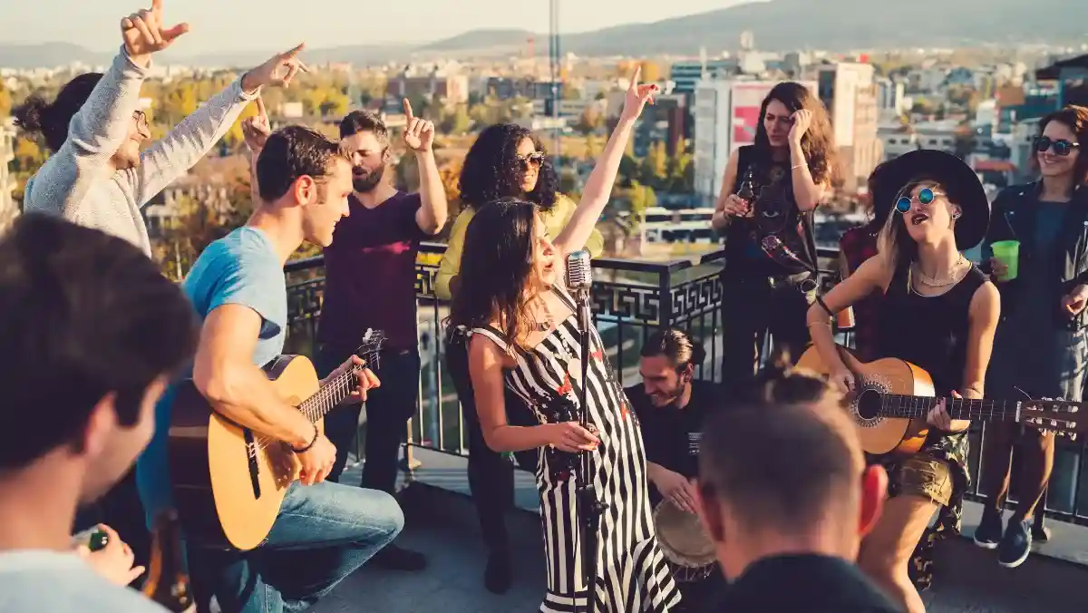 Group of friends enjoying a rooftop party, with people singing and playing guitars. Energetic atmosphere with a cityscape backdrop at sunset.
