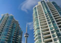 Two modern high-rise buildings with glass facades frame the CN Tower under a clear blue sky, creating an urban skyline scene in Toronto.