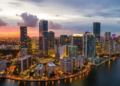 Aerial view of a vibrant city skyline at dusk, with illuminated skyscrapers reflecting on the water. A colorful sunset casts a warm glow over the urban landscape.