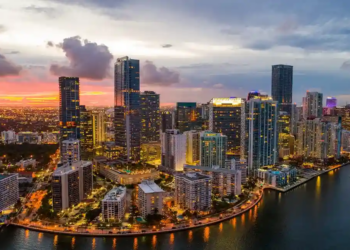 Aerial view of a vibrant city skyline at dusk, with illuminated skyscrapers reflecting on the water. A colorful sunset casts a warm glow over the urban landscape.