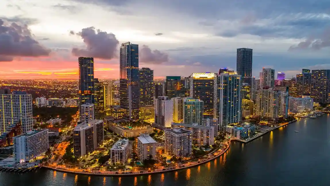 Aerial view of a vibrant city skyline at dusk, with illuminated skyscrapers reflecting on the water. A colorful sunset casts a warm glow over the urban landscape.