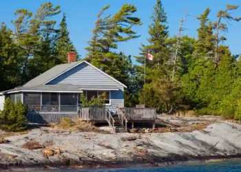 A cozy lakeside cabin with a wooden deck sits amidst lush greenery and tall pine trees. A Canadian flag waves beside the cabin under a clear blue sky.