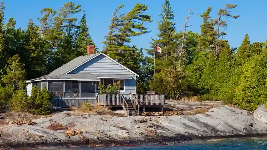 A cozy lakeside cabin with a wooden deck sits amidst lush greenery and tall pine trees. A Canadian flag waves beside the cabin under a clear blue sky.