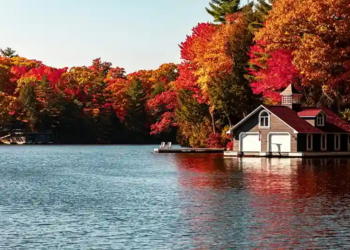 A serene lake scene with vibrant autumn foliage in red and orange hues. A quaint boathouse with a red roof sits by the water, reflecting a peaceful atmosphere.