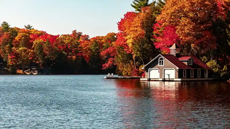 A serene lake scene with vibrant autumn foliage in red and orange hues. A quaint boathouse with a red roof sits by the water, reflecting a peaceful atmosphere.