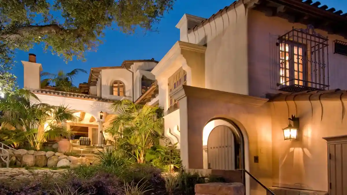 Luxurious Mediterranean-style villa at dusk, lit warmly with lanterns. Features arched doorways, ornate windows, and lush greenery creating an inviting ambiance.