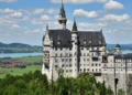 A fairytale-like castle stands majestically atop a lush, forested hill. Background features a serene lake and quaint village under a partly cloudy sky.