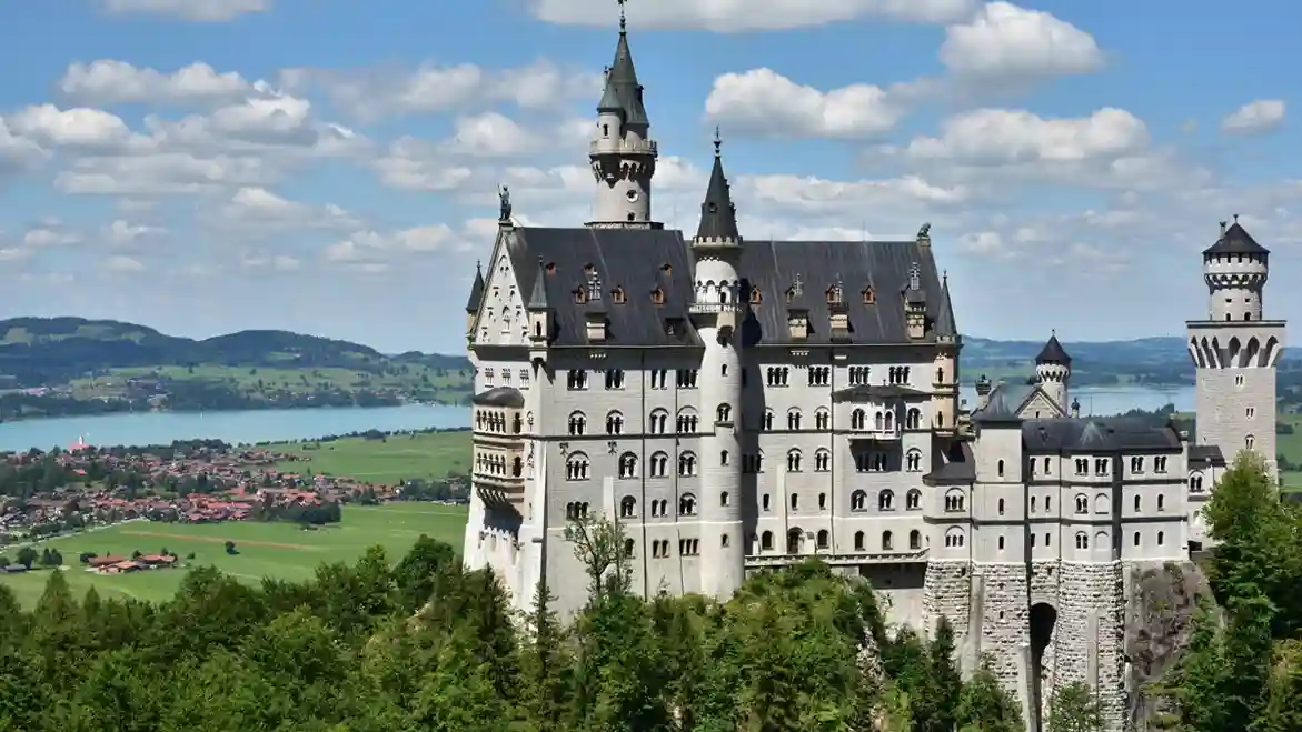 A fairytale-like castle stands majestically atop a lush, forested hill. Background features a serene lake and quaint village under a partly cloudy sky.