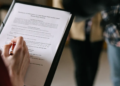 A person holding a pen fills out a form on a clipboard. The background shows two blurred figures, suggesting a professional or casual setting.
