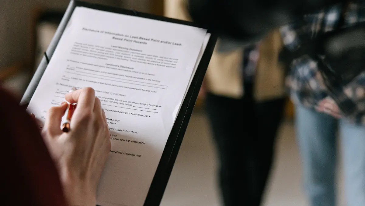 A person holding a pen fills out a form on a clipboard. The background shows two blurred figures, suggesting a professional or casual setting.