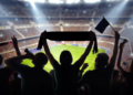 Silhouetted fans cheer in a packed, brightly lit stadium. One holds a scarf overhead, while others wave flags, creating an energetic, lively atmosphere.