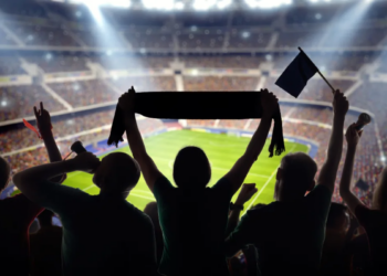 Silhouetted fans cheer in a packed, brightly lit stadium. One holds a scarf overhead, while others wave flags, creating an energetic, lively atmosphere.