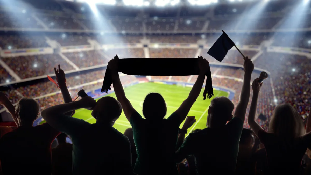 Silhouetted fans cheer in a packed, brightly lit stadium. One holds a scarf overhead, while others wave flags, creating an energetic, lively atmosphere.