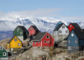 Colorful wooden houses scatter across a rocky hillside under a clear blue sky, with a snow-covered mountain range in the background, creating a serene, picturesque scene.