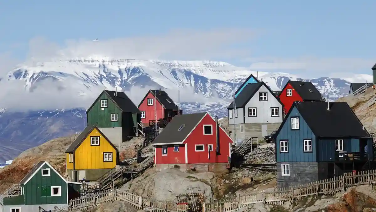 Colorful wooden houses scatter across a rocky hillside under a clear blue sky, with a snow-covered mountain range in the background, creating a serene, picturesque scene.