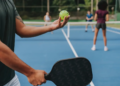 A person holds a pickleball paddle and ball, ready to serve on a blue court. Two players stand at the net in anticipation. The mood is focused and competitive.