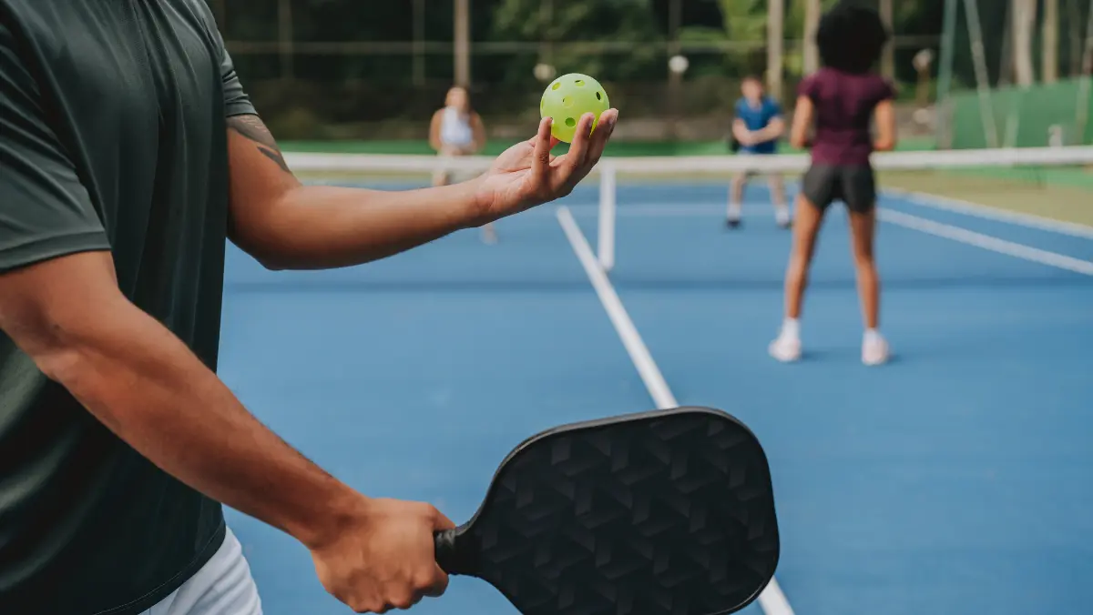 A person holds a pickleball paddle and ball, ready to serve on a blue court. Two players stand at the net in anticipation. The mood is focused and competitive.