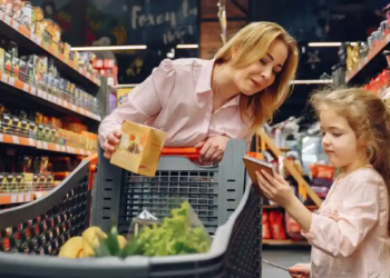 A woman and a child are grocery shopping in a brightly lit store aisle. The woman smiles at the child who is holding a packaging. A shopping cart is filled with groceries like bananas and greens, conveying a cheerful family outing.
