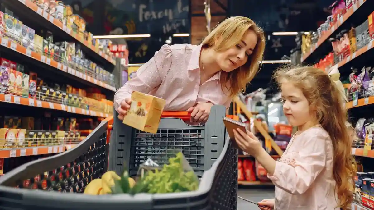 A woman and a child are grocery shopping in a brightly lit store aisle. The woman smiles at the child who is holding a packaging. A shopping cart is filled with groceries like bananas and greens, conveying a cheerful family outing.