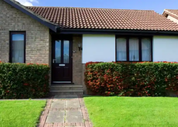 Brick bungalow with a red-tiled roof and a dark brown door, framed by hedges and a vibrant green lawn. The setting is peaceful and sunny.