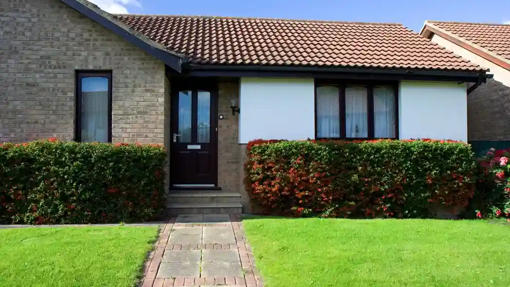 Brick bungalow with a red-tiled roof and a dark brown door, framed by hedges and a vibrant green lawn. The setting is peaceful and sunny.