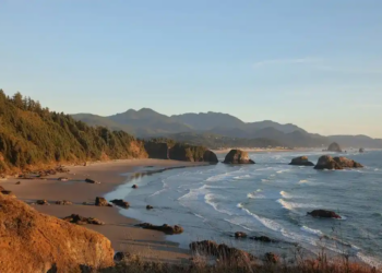 A tranquil coastal scene at sunset with waves lapping against a sandy shore, surrounded by rocky outcrops and forested cliffs under a clear sky.