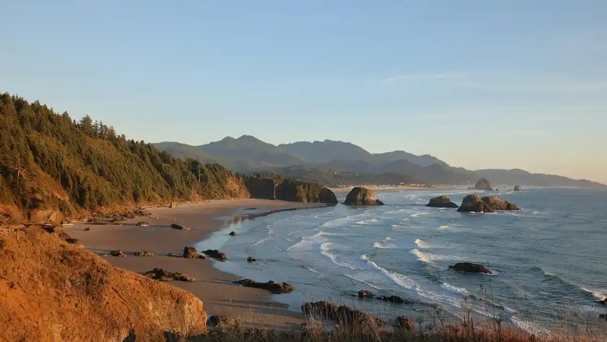 A tranquil coastal scene at sunset with waves lapping against a sandy shore, surrounded by rocky outcrops and forested cliffs under a clear sky.