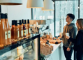 A young couple stands in a cozy café, holding coffee cups and browsing pastries. Warm lighting and a display of bottled drinks create a welcoming atmosphere.