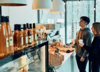 A young couple stands in a cozy café, holding coffee cups and browsing pastries. Warm lighting and a display of bottled drinks create a welcoming atmosphere.