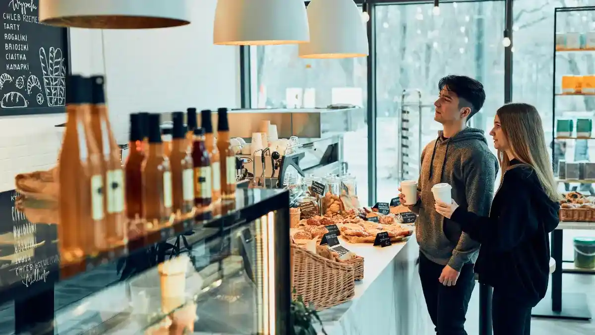 A young couple stands in a cozy café, holding coffee cups and browsing pastries. Warm lighting and a display of bottled drinks create a welcoming atmosphere.