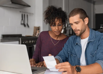 A couple sits at a kitchen table with a laptop, focused on reviewing a document. They appear thoughtful and engaged, conveying a collaborative tone.