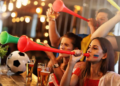 Fans in a lively bar blow colorful vuvuzelas, celebrating. A soccer ball and pints of beer are visible. The scene is energetic and joyful.