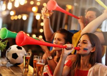 Fans in a lively bar blow colorful vuvuzelas, celebrating. A soccer ball and pints of beer are visible. The scene is energetic and joyful.