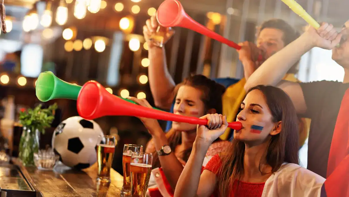 Fans in a lively bar blow colorful vuvuzelas, celebrating. A soccer ball and pints of beer are visible. The scene is energetic and joyful.