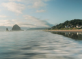 Calm ocean waves wash over a sandy beach with distant rocky formations and a lush, green hillside. A small coastal town is visible under a bright, clear sky.