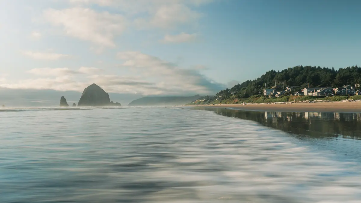 Calm ocean waves wash over a sandy beach with distant rocky formations and a lush, green hillside. A small coastal town is visible under a bright, clear sky.