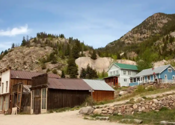 A rustic, small mountain village with vintage wooden buildings and colorful houses nestled against tree-covered hills under a clear blue sky.