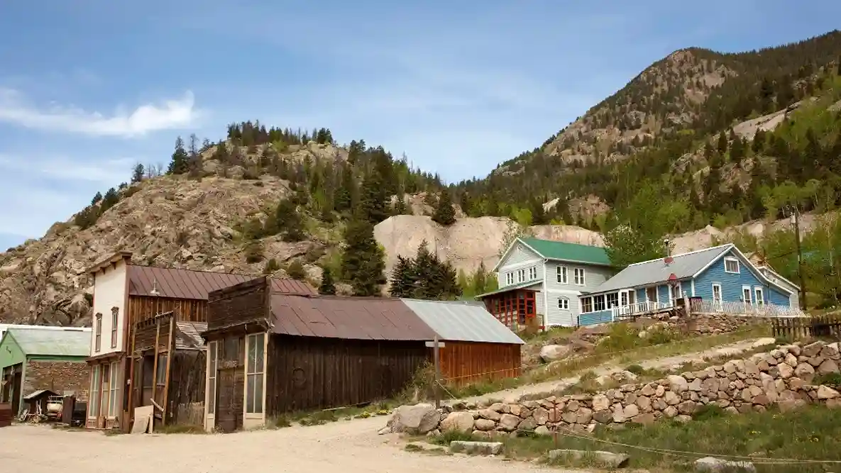 A rustic, small mountain village with vintage wooden buildings and colorful houses nestled against tree-covered hills under a clear blue sky.