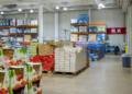 A spacious wholesale warehouse filled with stacked boxes of produce, drinks, and bulk goods on metal shelves. Bright lighting with an organized layout.