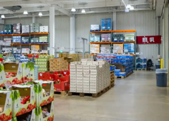 A spacious wholesale warehouse filled with stacked boxes of produce, drinks, and bulk goods on metal shelves. Bright lighting with an organized layout.