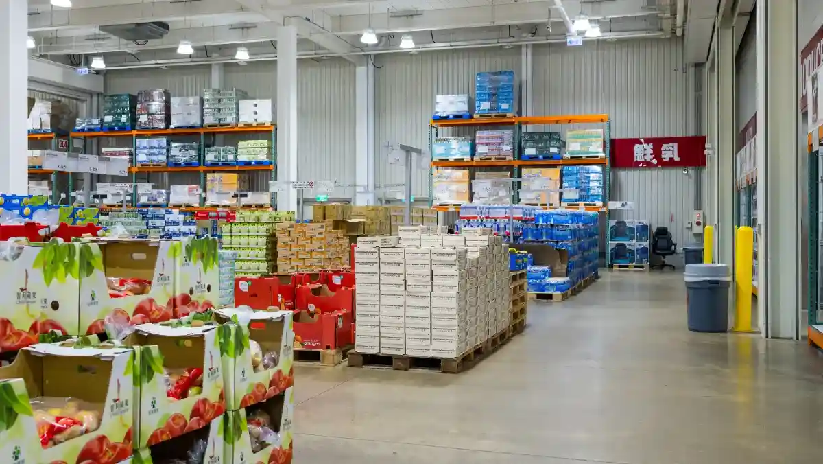 A spacious wholesale warehouse filled with stacked boxes of produce, drinks, and bulk goods on metal shelves. Bright lighting with an organized layout.