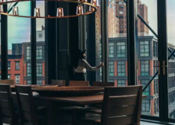 A modern dining area with a wooden table and chairs is set against large windows, offering a view of urban buildings. The ambiance is warm and cozy.