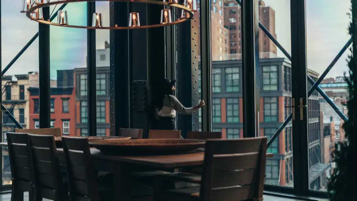 A modern dining area with a wooden table and chairs is set against large windows, offering a view of urban buildings. The ambiance is warm and cozy.